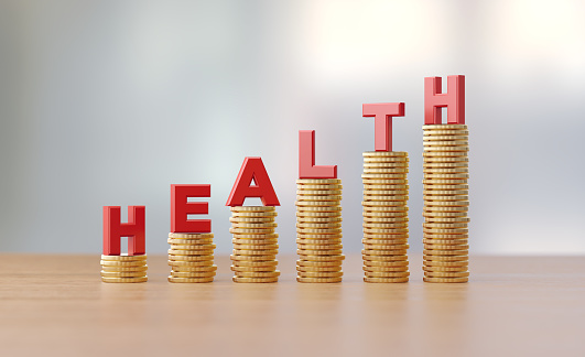 Red letters sitting over coin stacks write health before defocused background. Horizontal composition with copy space. Great use for finance and health concepts.