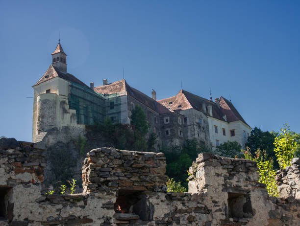 An old stone tower with small windows and a wooden shingle roof is nestled in a forest setting. The weathered walls and traditional roof design reflect the building's historical character, surrounded by lush green trees.