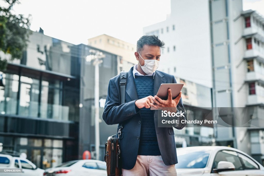 The road to business recovery begins now Shot of a masked businessman using a digital tablet against a city background Indian Ethnicity Stock Photo