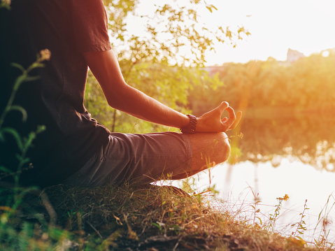 A young man in a lotus position on the bank of a river at sunset. Close-up