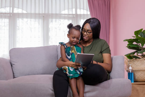 madre estaba enseñando a la hija a través de la tableta con alegría mientras estaba sentada en el sofá. negros o afroamericanos. concepto de estudio casero. concepto de padre-profesor. - child digital tablet mother teaching fotografías e imágenes de stock