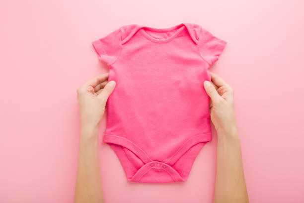 mains de mère de jeune adulte retenant le nouveau body de chéri sur le fond rose clair de table. couleur pastel. closeup. point de vue tiré. place vide pour le texte ou le logo sur les vêtements. vue de haut en bas. - onesie photos et images de collection