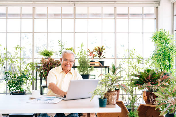 istock A senior retired old man dressed in casual style is using a laptop and smiling while sitting at home with indoor garden in the background. Retirement hobby and lifestyle. Grandfather work from home. 1294443757