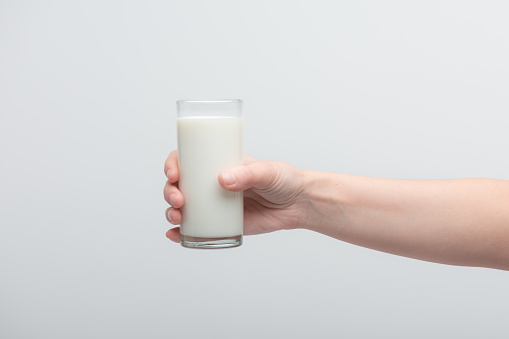 Naked female graceful hand holding transparent glass of milk over white background.