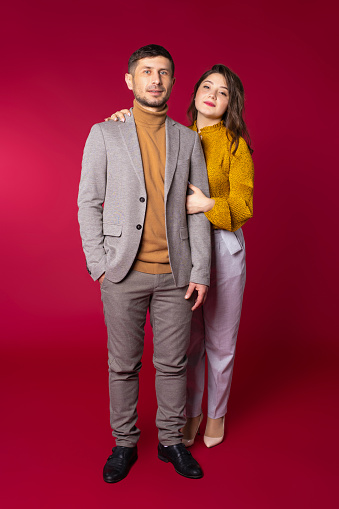 Full-length photo. Young loving couple with solid look wearing business clothes posing on red background in the studio. High quality photo