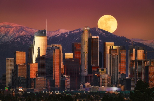 This photograph captures a full moon rising behind downtown Los Angeles' urban skyline and mountainous landscape.