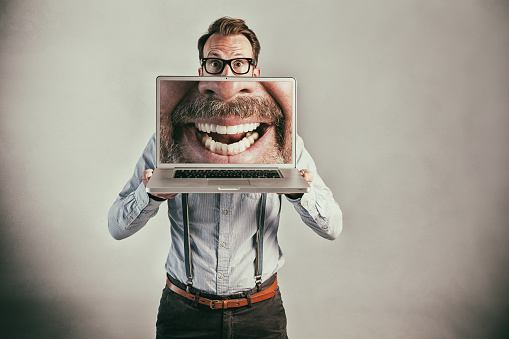Studio shot of a man holding a laptop screen in front of his face that displays a huge toothy grin