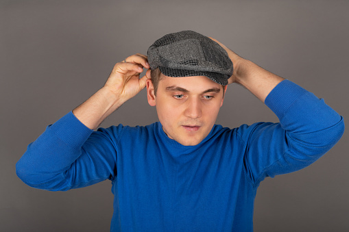Portrait of a young handsome caucasian man holding his cap in front of grey background