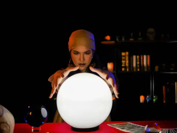 Asian woman fortune teller using magical occult divine power crystal call to forecast luck on red table.