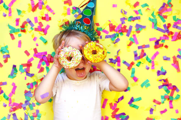 Little happy boy in birthday hat show tongue, having fun and holding two donuts near his eyes like glasses. Birthday concept stock photo