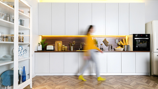 Blurred silhouette of young woman walking at home. Panoramic view of modern interior with white kitchen cupboards, wooden countertop, electric oven