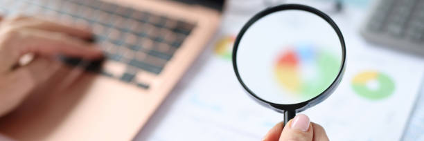 Woman holding magnifying glass in hands over documents with graphs and charts and working on laptop