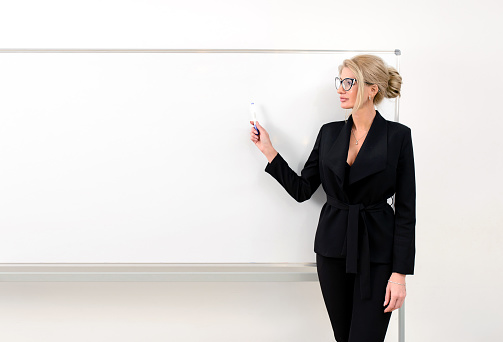 Teacher in a black suit near the whiteboard in a modern classroom points to the copy space. Isolated on white