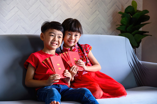 Portrait of young little Asian boy and girl smile happily and holding Red Envelopes (Angpow) containing money in hands as blessing from family, relatives and friends.