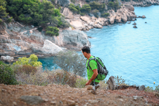High angle view of male hiker in 40s looking down at Mediterranean coastline while hiking on cliffside trail.