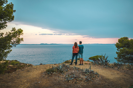 Rear view of male and female Spanish friends embracing while enjoying sunset on cliff overlooking Mediterranean Sea.
