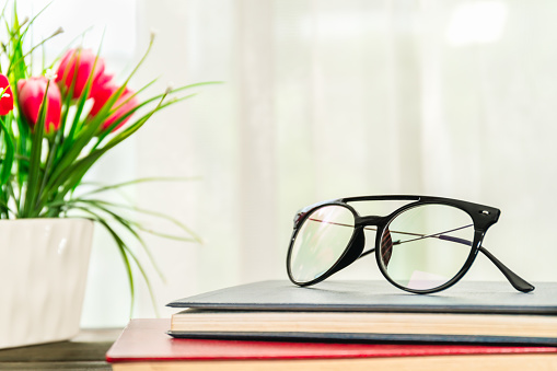 Reading glasses put on hardcover books over wooden table beside the window