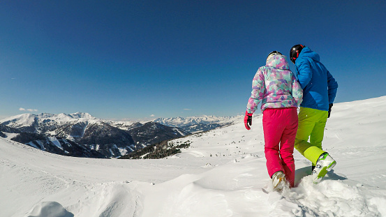 A couple in colorful skiing outfits and helmets walking on the fresh snow on top of Innerkrems, Austria. Everything is covered with fresh snow. High mountains in the back. Having fun and exploring