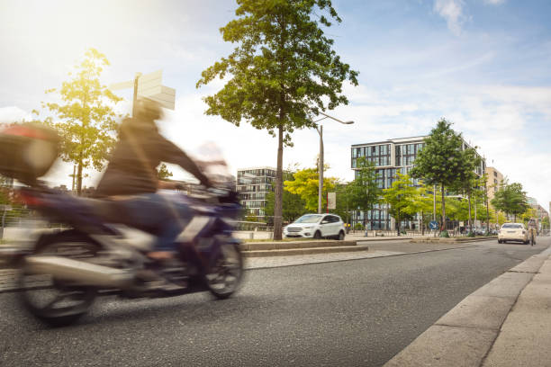 Public road in a German city. A blurred motorbike is driving by. Green trees, some cars and a blue sky in the background.