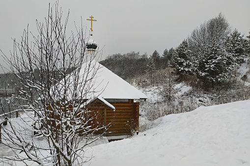 Small wooden building with golden orthodox cross against the background of winter forest and large snowdrifts.\