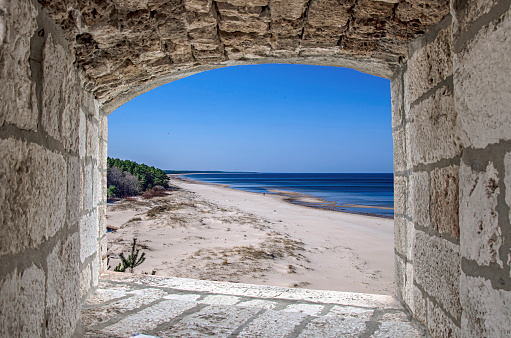 View from stone window of beach with white sand and blue sea/ocean. Beach with pine forest