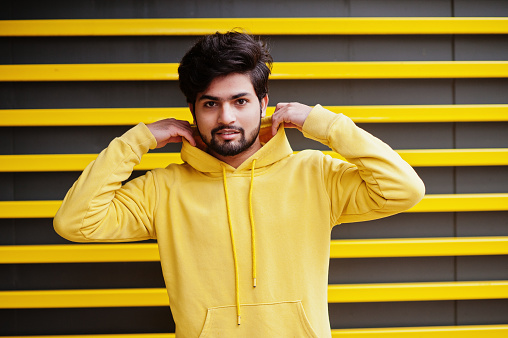 Urban young hipster indian man in a fashionable yellow sweatshirt. Cool south asian guy wear hoodie against striped background.