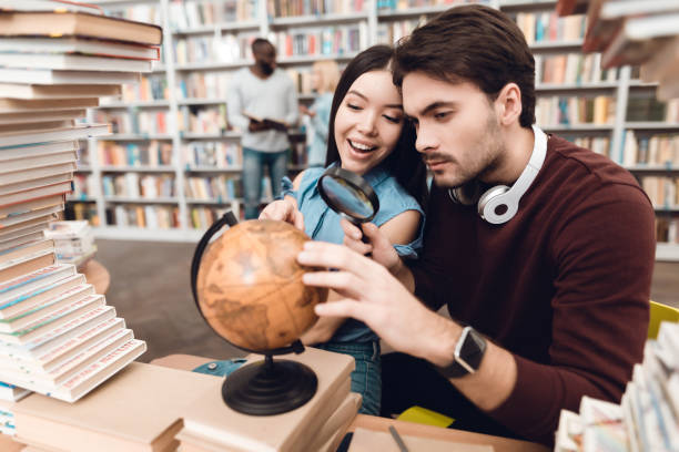 istock Ethnic asian girl and white guy surrounded by books in library. Students are using globe. 1294582365