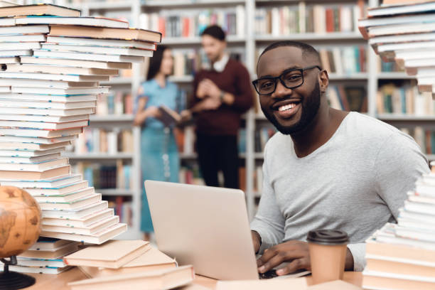 istock Ethnic african american guy surrounded by books in library. Student is using laptop and drinking coffee. 1294582430