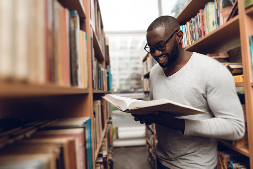 A man looks in a book and smiles in the library. Black man reading a book while standing in a large library.