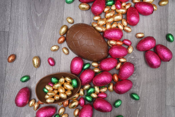 istock Pile or group of multi colored and different sizes of colourful foil wrapped chocolate easter eggs in pink, red, gold and green. Large halves of a brown milk chocolate egg have mini eggs inside, on a grey wooden background. 1294583379