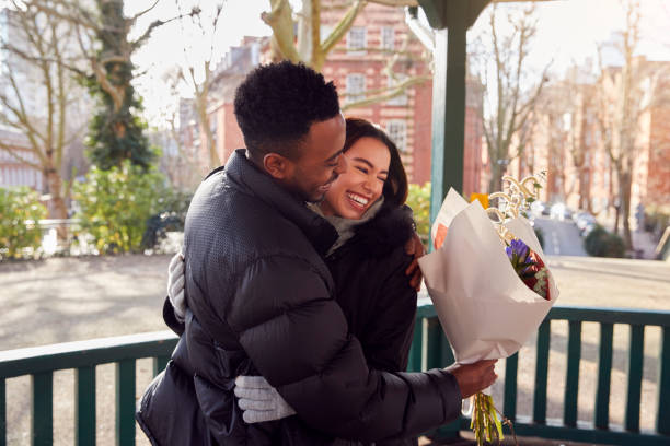 romantic man hugging young woman after giving her bouquet of flowers as they meet in city park - men african descent giving flower imagens e fotografias de stock