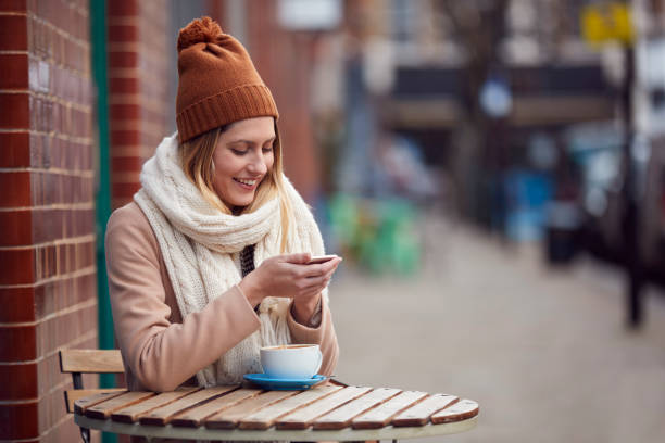 weibliche kundin sendet textnachricht sitzen vor coffee shop auf belebte stadt high street im herbst - autumn women clothing retail stock-fotos und bilder