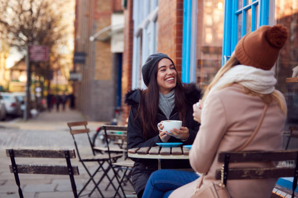 dos amigas femeninas se reúnen sentadas fuera de la cafetería en la calle principal de la ciudad - outdoors drinking women friendship fotografías e imágenes de stock