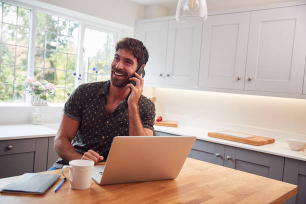 istock Man In Kitchen With Mobile Phone Working From Home Using Laptop During Health Pandemic 1294587390