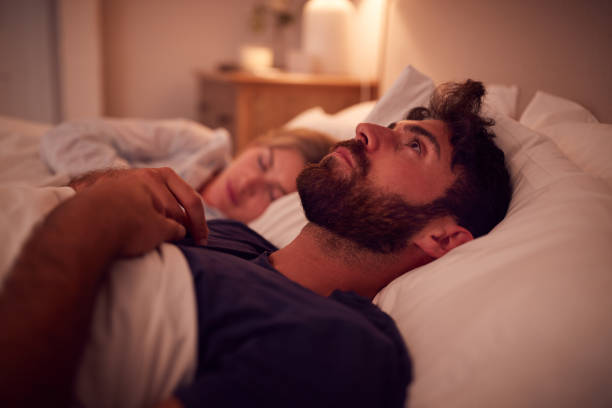 coppia con uomo sdraiato a letto sveglio di notte soffrendo di insonnia - insonnia foto e immagini stock