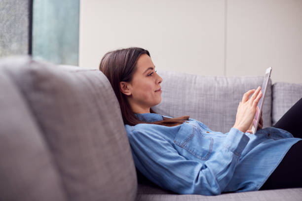 istock Smiling Young Woman At Home Lying On Sofa Looking At Digital Tablet 1294588002