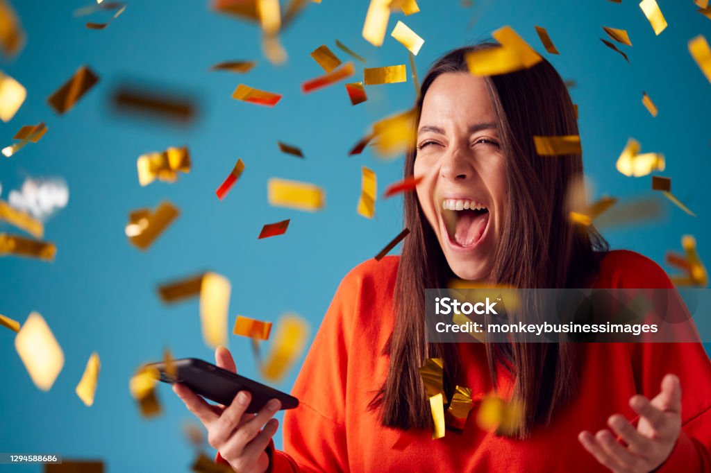 Celebrating Young Woman With Mobile Phone Winning Prize And Showered With Gold Confetti In Studio Winning Stock Photo