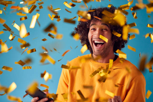 celebrando al joven con el premio ganador del teléfono móvil y duchado con confeti de oro en el estudio - ganar fotografías e imágenes de stock