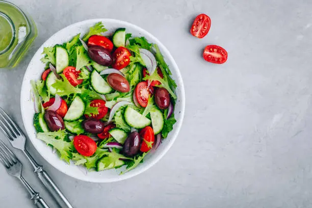 Healthy green salad with fresh tomato, cucumber, red onion, olives and lettuce in bowl on gray stone background. Top view, copy space.