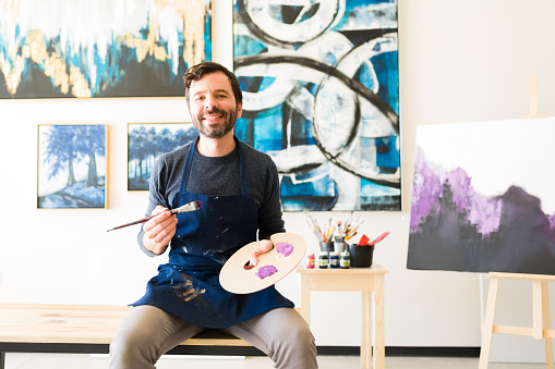 Portrait of a happy male painter in his 30s standing in his art studio with a brush and a palette. Caucasian man looking proud next to his new unfinished canvas
