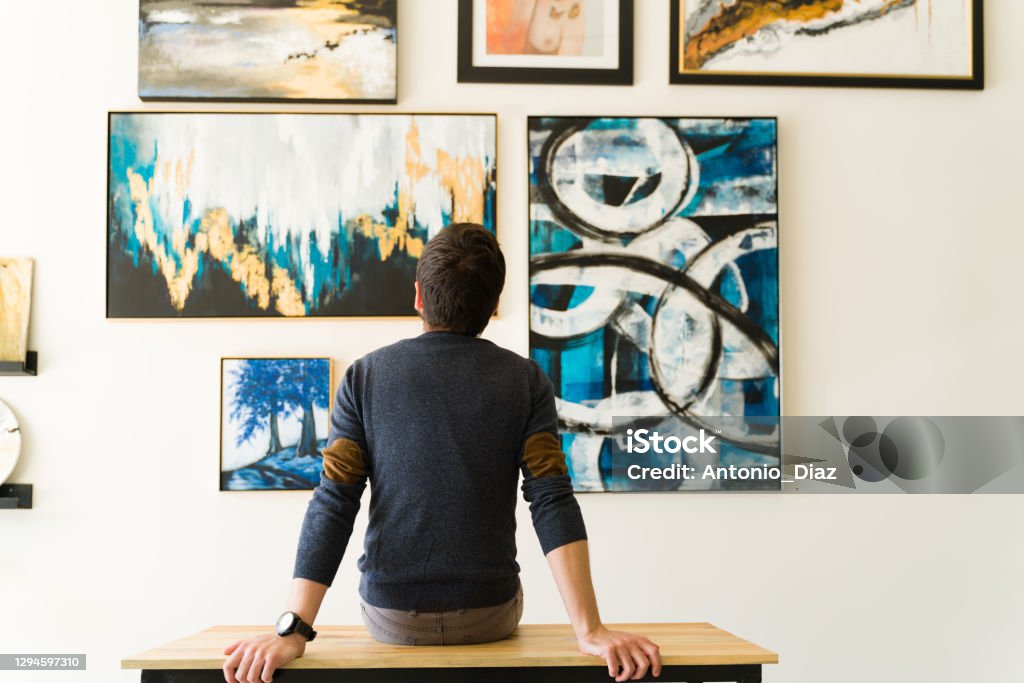 Rear view of a guy in his 30s looking at an art exhibition Hispanic male visitor looking reflective while sitting on a bench and admiring the various paintings on the wall of an art gallery Art Stock Photo