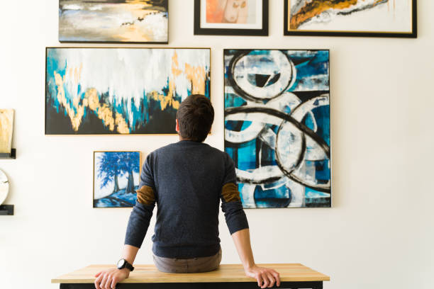 Rear view of a guy in his 30s looking at an art exhibition Hispanic male visitor looking reflective while sitting on a bench and admiring the various paintings on the wall of an art gallery art stock pictures, royalty-free photos & images