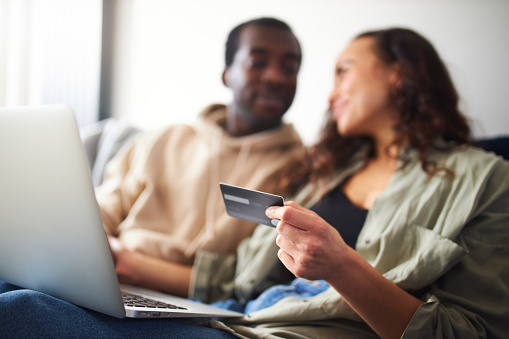 Young mixed ethnicity couple sit side by side on sofa at home holding credit card and shopping online using laptop with focus on foreground