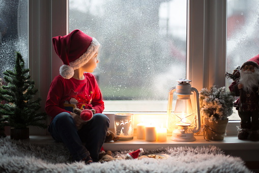 Beautiful toddler child, boy, waiting on the window on Christmas eve, looking for Santa Claus curiously