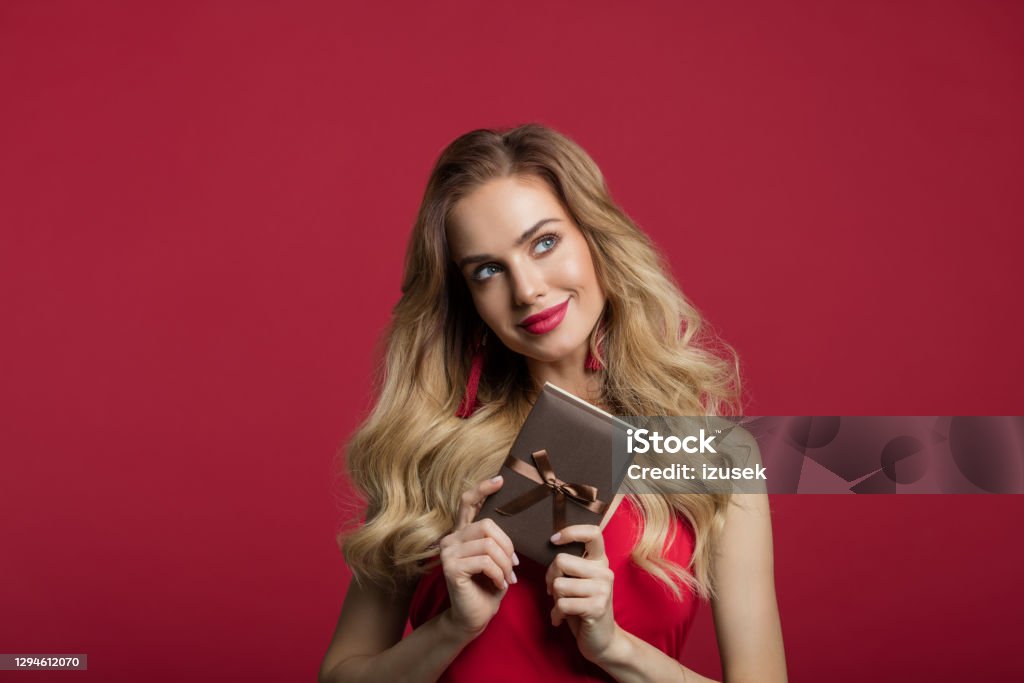 Beautiful woman holding gift box Portrait of beautiful long blond hair woman wearing red dress holding gift box and looking away. Studio shot against red background. Valentine’s day concept. One Woman Only Stock Photo