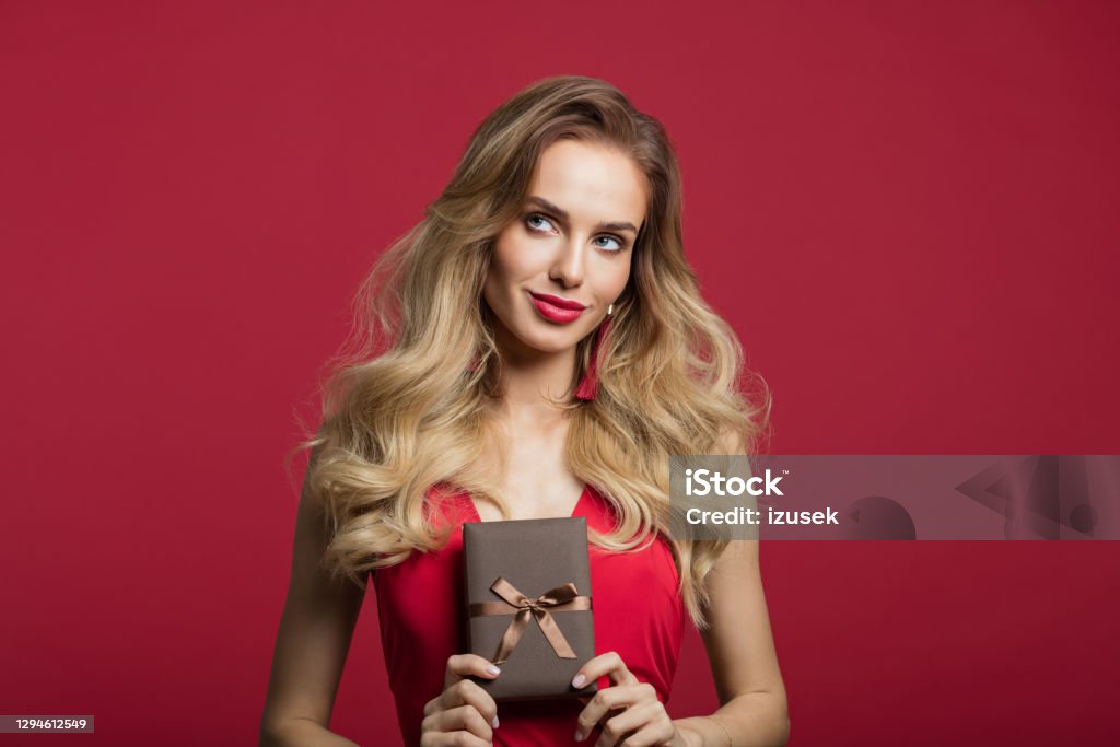 Beautiful woman holding gift box Portrait of beautiful long blond hair woman wearing red dress holding gift box and looking away. Studio shot against red background. Valentine’s day concept. Women Stock Photo