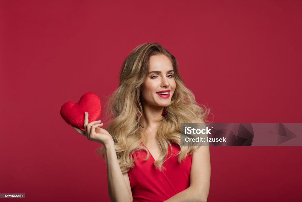 Beautiful woman holding red heart Portrait of beautiful long blond hair woman wearing red dress holding red heart and looking at camera. Studio shot against red background. Valentine’s day concept. One Woman Only Stock Photo