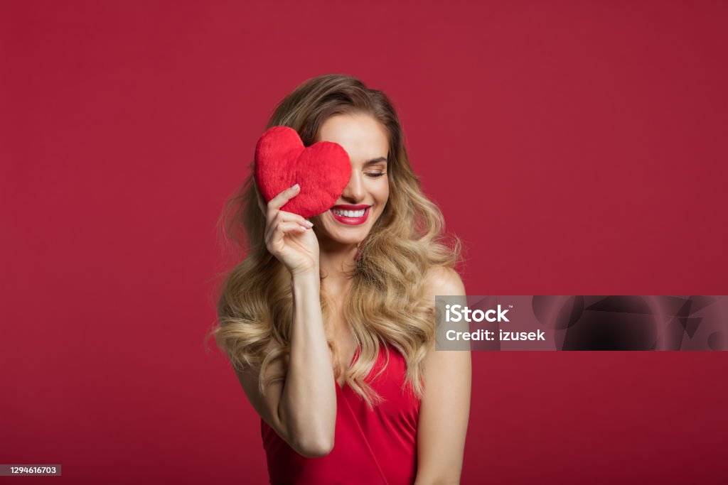 Happy woman holding red heart Portrait of beautiful long blond hair woman wearing red dress covering eye with red heart. Studio shot against red background. Valentine’s day concept. Valentine's Day - Holiday Stock Photo