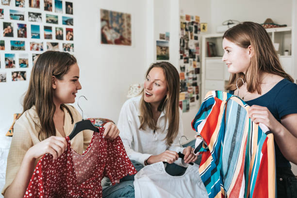 three young woman showing clothes from hatstand to eachother young woman presenting a dress on coat hanger to her female room mates indoors preppy fashion stock pictures, royalty-free photos & images