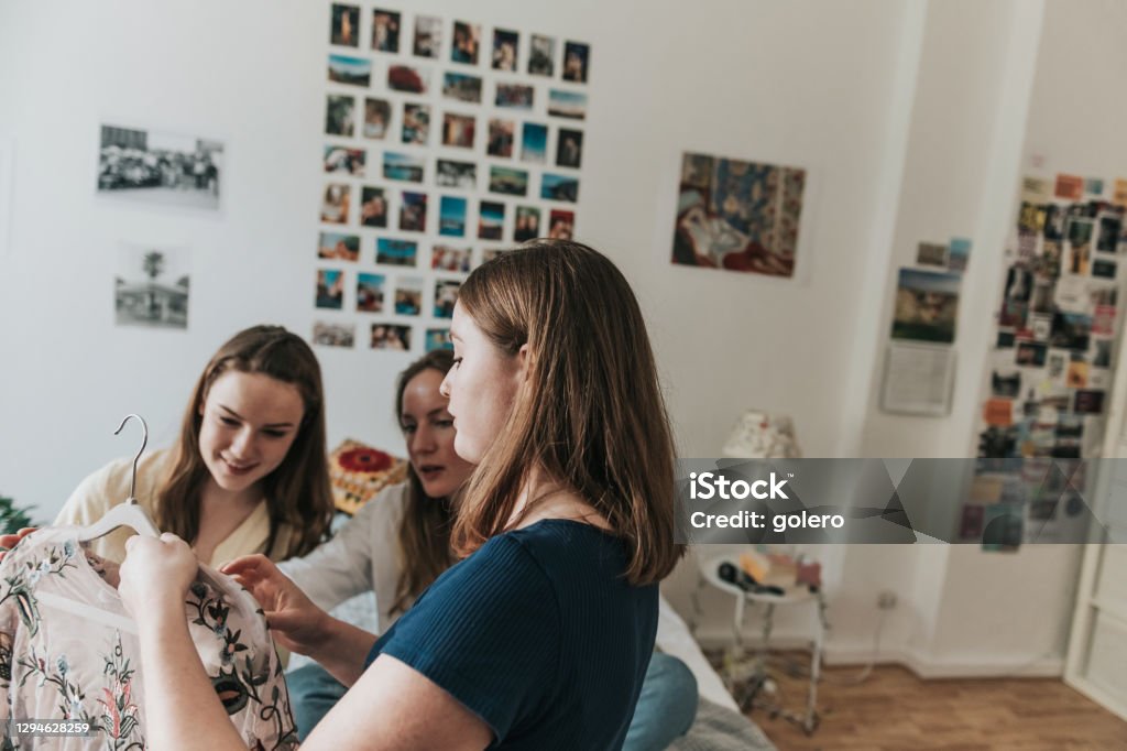 three young woman showing clothes from hatstand to eachother young woman presenting a dress on coathanger to her female room mates indoors Choosing Stock Photo